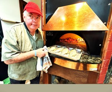 A man in a red cap bakes fish in a wood-fired oven, holding a tray with several fish ready to be cooked.