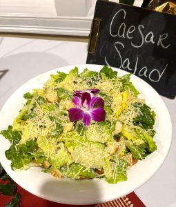 A bowl of Caesar salad topped with croutons, grated cheese, and a purple edible flower, next to a sign labeled "Caesar Salad.