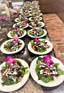 Multiple plates of salad with greens, tomatoes, dressing, and purple orchids arranged in rows on a granite countertop.
