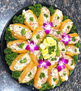 A platter of lobster rolls garnished with parsley, lime wedges, and purple orchids on a dark granite countertop.