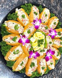 A platter of lobster rolls garnished with parsley, lime wedges, and purple orchids on a dark granite countertop.