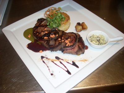 Grilled steak with sauce, vegetables, mashed potatoes, and a side of herb butter on a square white plate.