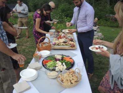 People gather around an outdoor buffet table with vegetables, dips, bread, and assorted appetizers on white tablecloths.