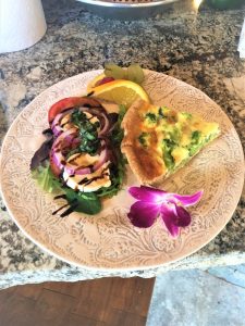 A plate with a slice of broccoli quiche, mixed greens with tomato and onion, orange slice, grapes, and a purple flower.