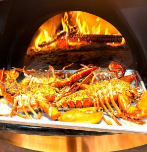 Lobsters roasting on a baking tray inside a wood-fired oven with flames visible in the background.